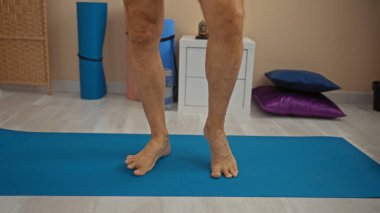 Sağlıklı bir odada mavi yoga minderinin üzerinde duran bir adam renkli yastıklar ve egzersiz aletleriyle rahatlama ve dayanıklılığı ima ediyor..