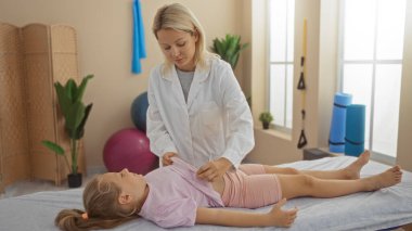 Kadın terapist, arka planda egzersiz ekipmanıyla, parlak bir klinikte tedavi masasında çocuk hasta değerlendirmesi yapıyor.