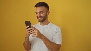 Akıllı telefon kullanırken gülümseyen sakallı genç İspanyol bir adam, sıradan bir tişört portresinin içinde canlı sarı bir arkaplan karşısında duruyor..