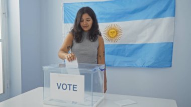 Arka planda Arjantin bayrağı olan kapalı bir seçim odasında oy veren genç bir kadın, oy pusulasını şeffaf bir kutuya atıyor..