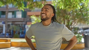 Saçları örgülü gülümseyen Afrikalı Amerikalı genç adam, açık havada rahat ve iyimser bir havayı somutlaştıran, güneş ışığıyla yıkanmış bir şehir parkında kendinden emin bir şekilde duruyor.