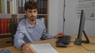 Ofisteki bir masada çalışan sakallı genç adam, kitaplarla ve dosyalarla dolu rafların arasında bilgisayarın önündeki belgelere odaklanıyor..