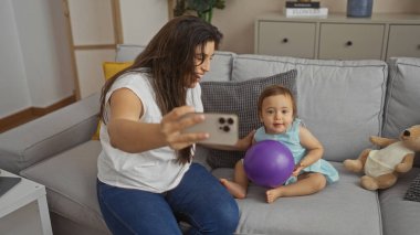 Bebek ile selfie çeken bir kadın oturma odasında kanepeye oturmuş aile zamanı ve sevgi gösterisi yapıyor.