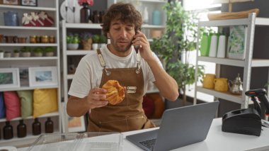 Elinde pasta tutan ve dekore edilmiş bir mağazanın içinde telefonla konuşan genç bir adam arkasında çeşitli dekorasyon malzemeleriyle bir bilgisayarın başında çalışıyor..