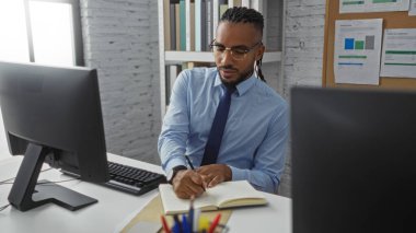 Örgülü genç adam modern ofis ortamında bilgisayar ve belgelerle birlikte deftere yazı yazıyor. Kapalı alanda odaklanmış ve profesyonel bir atmosfer yaratıyor.