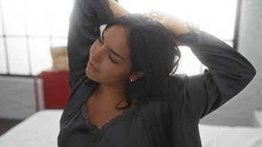 Genç bir kadın, büyük pencereli, parlak, rahat bir yatak odasında siyah pijama giyerek yatakta geriniyor.