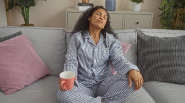 Huzurlu ve sade bir atmosfer yaratan, minder ve bitkilerle çevrili, rahat bir oturma odasında kanepede otururken, çizgili pijama giymiş bir kadın..