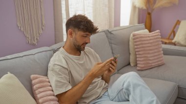 Kafkasyalı genç bir adam evdeki koltukta dinleniyor. Telefonunu evin içinde rahat bir oturma odasında kullanıyor.