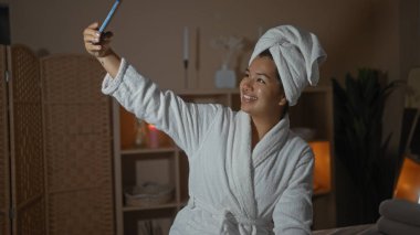 Güzel, genç bir İspanyol kadın spa 'da dinlenirken selfie çekiyor. Beyaz bir bornoz ve kafasında havluyla kapalı bir sağlık merkezinde..