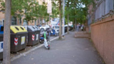 Yoğun bir şehir ortamında, ağaçların yol boyunca dizildiği ve arka planda insanların bulunduğu bulanık çöp konteynırları ve scooterların yer aldığı kentsel alandan arındırılmış sokak manzarası.