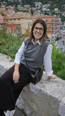 Kadın taştan bir duvarda oturuyor, arkasında renkli bir villefranche, Fransa binaları ve deniz var..