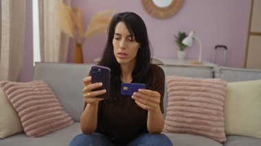 Genç, çekici, İspanyol bir kadın elinde akıllı telefon ve kredi kartıyla oturma odasında oturuyor. Modern bir evin içinde düşünceli görünüyor..