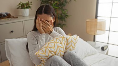 Yatak odasında oturan genç bir kadın dekoratif bir yastık tutarken elleriyle yüzünü örtüyor. Ev dekorasyonu ve hafif ışıklandırmayla çevrili.