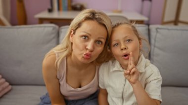 Sarışın anne ve kızı, sıcak bir oturma odasındaki kanepede birlikte komik suratlar yapıyor.