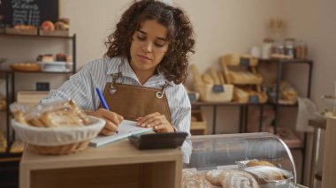 Fırında çalışan bir kadın, bir kalem ve not defteriyle ekmek dolu bir sepetin yanına notlar yazıyor, etrafı fırınlanmış mallarla çevrili.