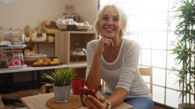 Fırında gülümseyen sarışın bir kadın telefon kullanırken, arka planda bir bitki ve hamur işleriyle bir masanın önünde oturuyor..