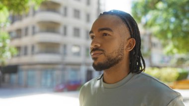 Saçlı genç adam güneşli şehir parkında yürüyor. Etrafında binalar var. Düşünceli bir ifade ve şehir manzarasına karşı şık bir görünüm sergiliyor..