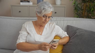 Gri saçlı, gözlüklü ve beyaz gömlekli yaşlı bir kadın modern iç dekorlu bir oturma odasında akıllı telefon kullanıyor..