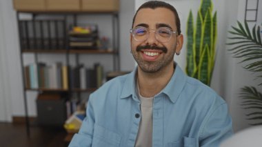 Ofis ortamında gülümseyen genç, İspanyol bir adam. Arkasında raflar ve bitkiler var..