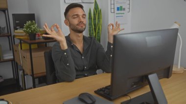 Sakallı genç bir İspanyol adam ofisteki masasında meditasyon yapıyor. İçerde sakin bir ortam yaratıyor, işyerinde stres atağı vurguluyor..