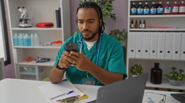 Klinikte saçı örgülü genç bir adam hastane odasında çevresi tıbbi ekipmanlarla çevrili telefonları kontrol ediyor ve yeşil önlük giyiyor..