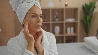 Kafasında havlu olan olgun bir İspanyol kadın huzurlu bir spa ortamında rahatlatıcı bir anın tadını çıkarıyor, bir sağlık odasında yüzüne nazikçe dokunuyor..