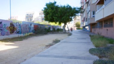 Blurred background outdoors showing a defocused walkway with colorful graffiti, surrounded by buildings and trees in a sunny urban setting. clipart