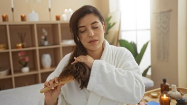 Esmer saçlarını spa odasında tarayan bir kadın, arka planda parlayan mumlarla güzelliğini artırıyor. Huzurlu bir iç güzellik salonu ortamında..