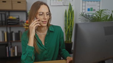 Gözlüklü ve yeşil tişörtlü genç çekici bir kadın, arka planda bitki ve kitap rafları olan modern bir ofis odasında bilgisayarının başında çalışırken telefonla konuşuyor..