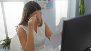 Ofisindeki belgeleri inceleyen genç bir kadın gözlüklü ve modern bir iş ortamında ciddi bir ifadeyle stresli görünüyor.