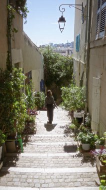 Woman exploring narrow european alley surrounded by lush plants on sunny day in charming cityscape with old lanterns casting shadows and historic stone path. clipart