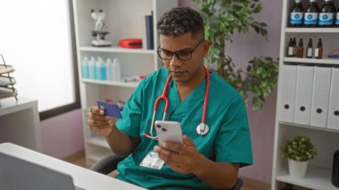 Yeşil önlüklü İspanyol erkek hemşire, arka planda tıbbi malzemeler bulunan modern bir klinik işyerinde özenle çalışarak bir hastane odasında akıllı telefon ve kredi kartı kullanıyor..