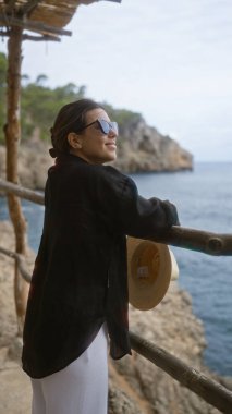 Genç bir kadın Mallorca, İspanya 'daki Cala Deia' da manzaranın tadını çıkarıyor. Güneş gözlüğü takıyor ve elinde hasır bir şapkayla açık havada ahşap korkuluklara yaslanıyor.