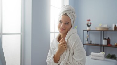 Havluya sarılmış bir kaplıcada dinlenen bir kadın, parlak, huzurlu bir sağlık merkezinde sakin bir ifadeyle gülümsüyor arka planda raflar ve büyük pencerelerden süzülen güneş ışığı