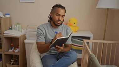 Örgülü genç bir adam, bebek beşiğinin yanındaki rahat bir yatak odasında, iç mekanda bebek eşyalarıyla dolu bir not defterine yazı yazıyor..