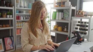 Ev dekorasyonu mağazasında dizüstü bilgisayarda çalışan genç bir kadın. Sarı saçlı ve gözlüklü. Etrafı çeşitli dekoratif eşyalar ve raflarda iç mekan bitkileriyle çevrili.