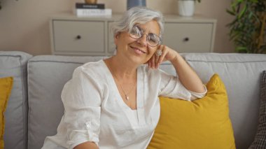 Gri saçlı, beyaz, olgun, kadın, gülümseyen, evdeki bir kanepede dinlenen, oturma odası, gözlük takan, arka planda sarı yastıklar olan beyaz bir gömlek..