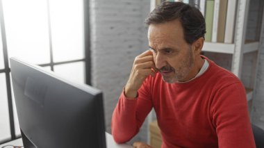 Kırmızı kazaklı İspanyol bir adam bir ofis ortamında bilgisayar ekranına odaklanmış düşünceli bir konsantrasyon ve işyeri ortamı vurguluyor..