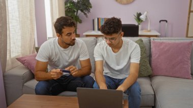 Baba ve oğul rahat bir oturma odasında dizüstü bilgisayar kullanarak aile sevgisini ve aile bağlarını modern dekor ve günlük kıyafetlerle sergiliyorlar.