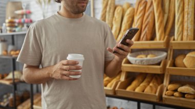 Fırında kahve fincanı ve akıllı telefon tutan genç adam taze ekmek raflarıyla çevrili..