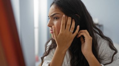 Kaplıcadaki aynada yüzünü inceleyen bir kadın. Düşünceli bir ifade sergiliyor. Kapalı mekan ışıklarıyla..