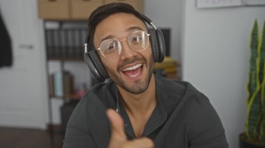 Kulaklık ve gözlük takan, gülümseyen ve ofis ortamında onaylayan sakallı genç İspanyol bir adam..
