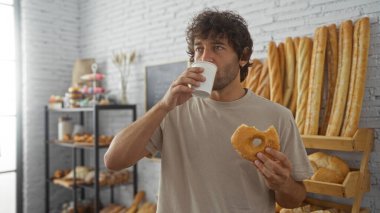 Handsome young man drinking coffee and eating a donut while standing in a cozy bakery with loaves of bread and pastries in the background clipart