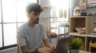 Sıcak fırında ya da kahve dükkanında dizüstü bilgisayarda çalışan genç İspanyol bir adam, çeşitli fırınlanmış mallarla dolu raflarla çevrili, günlük giysiler giyiyor ve odaklanmış bir ifade sergiliyor.