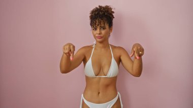 Beyaz bikinili genç bir kadın pembe arka planda dikilirken aşağıya doğru bakıyor..