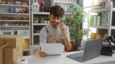 Ev dekorasyon dükkanında çalışan sakallı genç bir adam, telefonla konuşuyor, evrak işleri yapıyor, önlük giyiyor, dizüstü bilgisayara bakıyor, kutular, bitkiler ve dekorasyon malzemeleriyle çevrili..