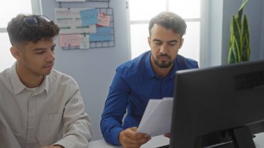 Bir ofiste birlikte çalışan iki İspanyol adam, belgeler ve bilgisayar ekranlarına odaklanmış, profesyonel çevreyi takım çalışması ve işbirliği ile somutlaştırıyorlar..