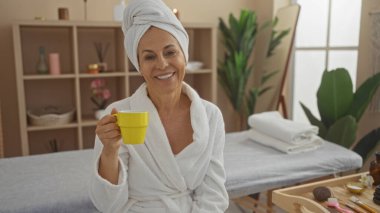 Kaplıcada çay içen bir kadın, beyaz bir bornoz ve türban giyiyor, rahat bir sağlık merkezinde arka planda bitkilerle oturuyor, gülümsüyor ve rahatlıyor.