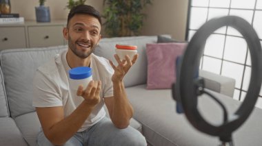Genç bir İspanyol, elinde iki krema kavanozu tutarken ve rahat bir dairede video çekerken gülümserken.