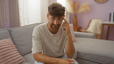 Genç bir adam, modern bir dekorla oturma odasında oturuyor, gülümsüyor ve şakağını işaret ediyor, arka planda rahat bir kanepesi olan sıradan bir tişört giyiyor.